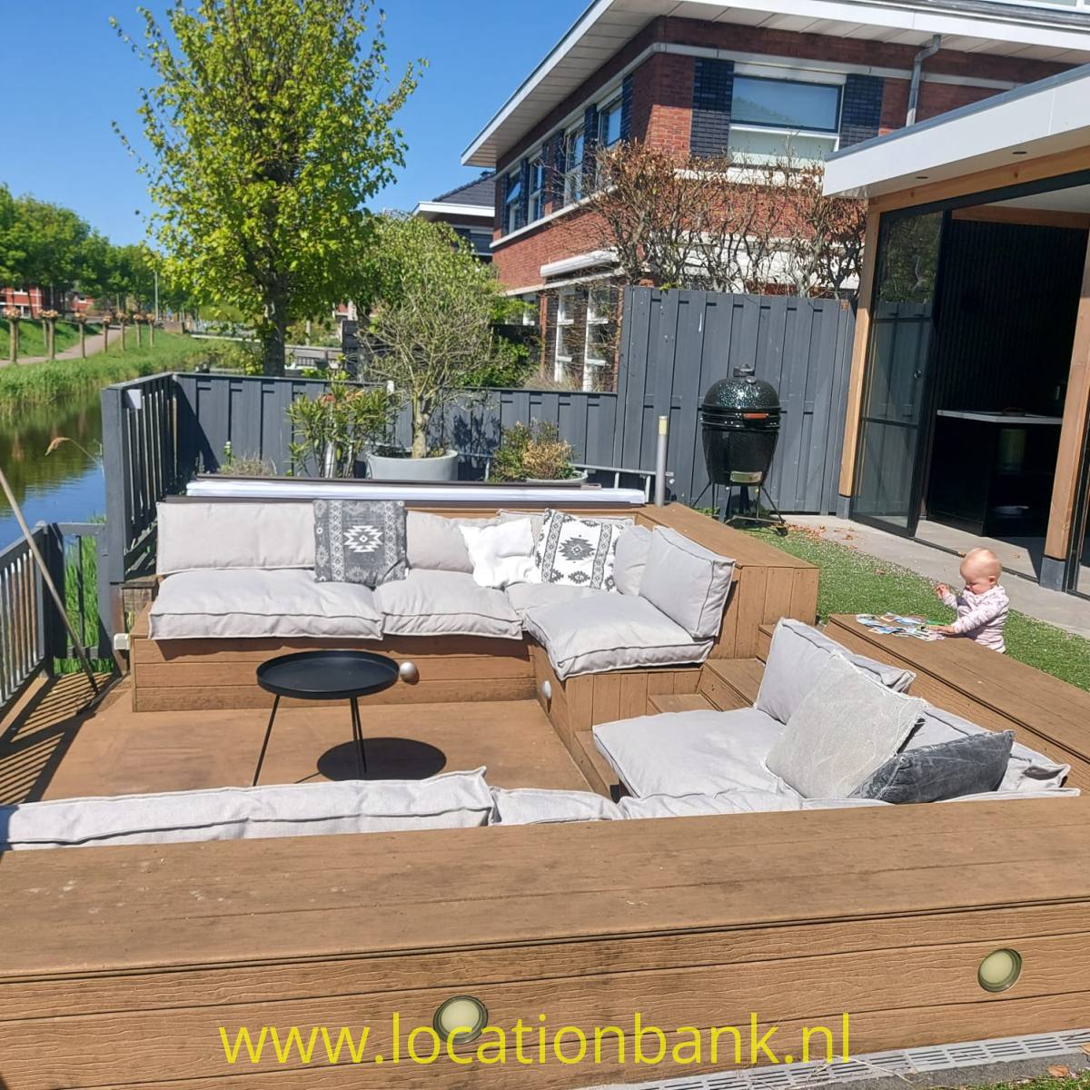 Holzterrasse am wasser mit einer Sitzgruppe, Pflanzgefäßen, einem Grill und Blick auf einen schmalen Kanal neben einem Haus.