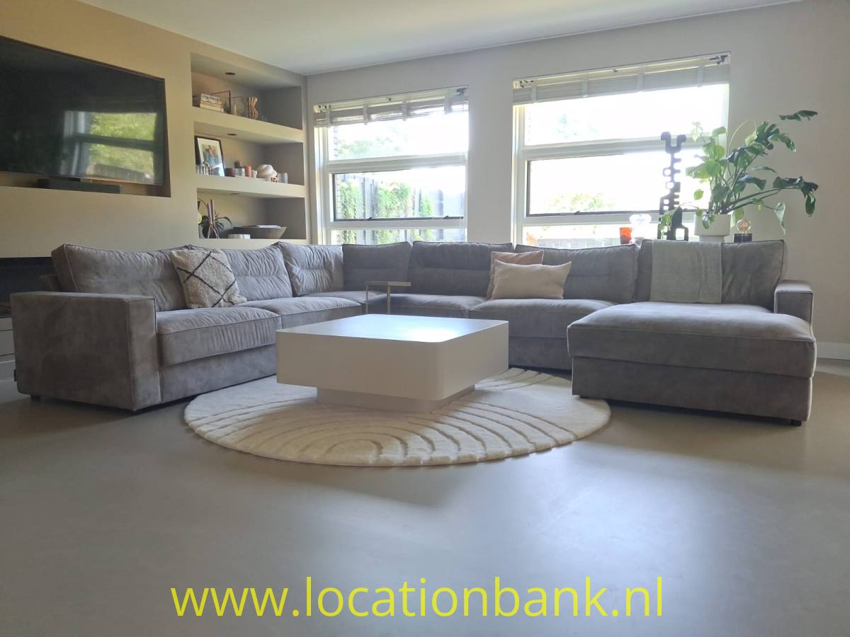 Graues Ecksofa mit Longchair, niedrigem weißem Couchtisch und rundem Teppich vor hohen Fenstern.