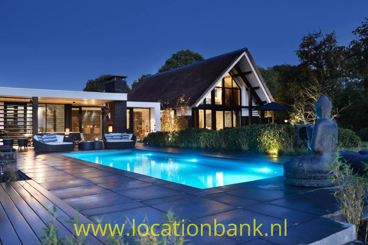 Reetgedecktes Haus mit verglaster Giebelfassade, beleuchtetem Swimmingpool, Terrasse und Skulptur in der Dämmerung.