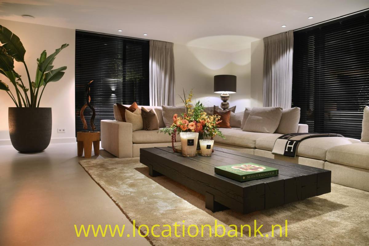 Wohnzimmer mit einem beigen Ecksofa, einem schwarzen Couchtisch, einer großen Zimmerpflanze und Fenstern mit dunklen Jalousien.