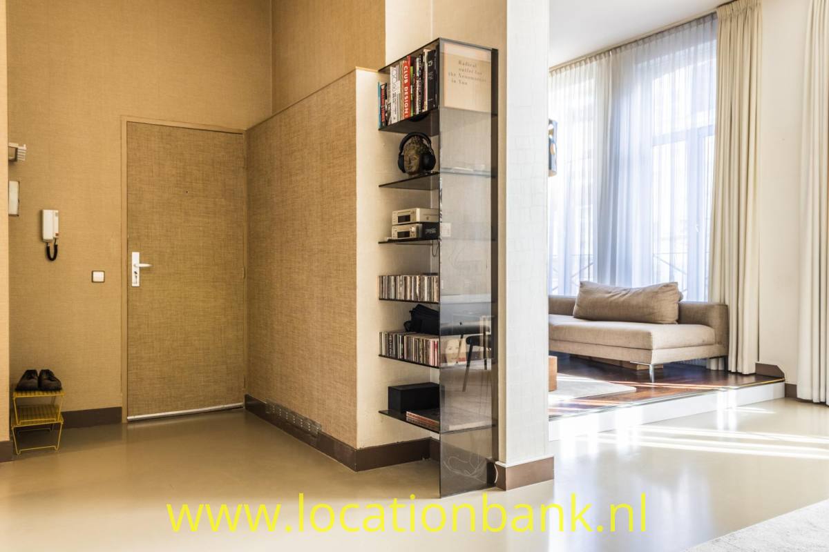Ein Flur mit einem gläsernen Bücherregal bietet durch einen offenen Durchgang den Blick auf ein Wohnzimmer mit hohen Fenstern.