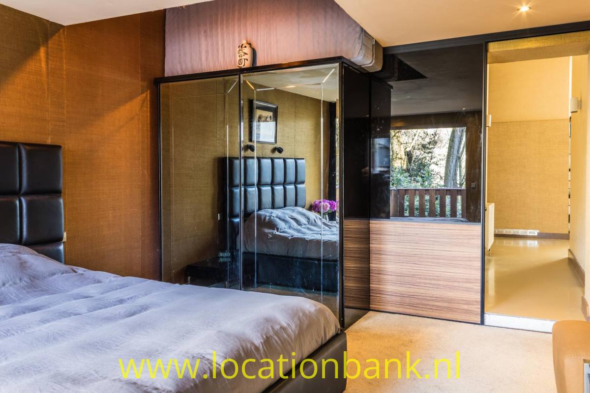 Schlafzimmer mit einem großen Bett, einem verglasten Kleiderschrank und einem offenen Durchgang zu einem Balkon.