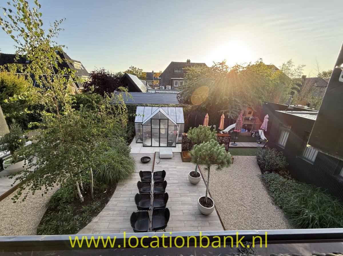 Eine Terrasse zeigt ein gläsernes Gewächshaus, einen langen Tisch mit Stühlen, Pflanzgefäße und eine Kiesfläche.