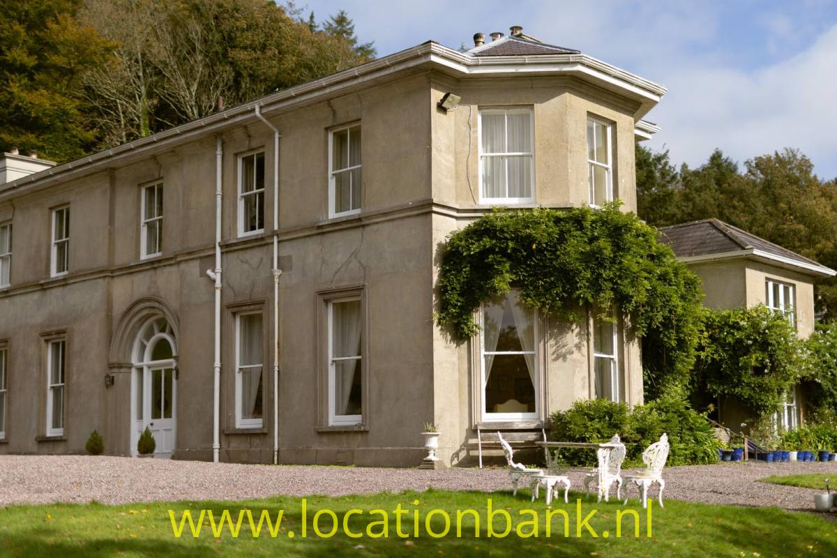 Helles zweigeschossiges Landhaus mit einer von Efeu bedeckten Fassade, einem rundbogigen Eingang und weißen Stühlen auf einer Kiesterrasse.