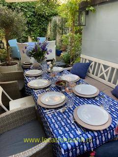 Gedeckter Tisch im Freien mit blau gemusterter Tischdecke, Geschirr, Gläsern und Blumen, umgeben von Stühlen, Pflanzen und einer weißen Wand.