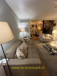 Wohnzimmer mit einem beigen Ecksofa, einer Stehlampe, einem Sessel, einem Couchtisch und einer Treppe im Hintergrund.