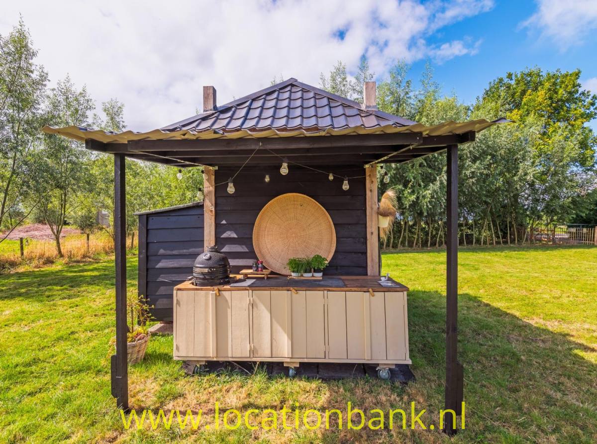 Ein hölzerner Stand mit Ziegeldach, Theke, Lichterkette und großer runder Wanddekoration steht auf einer Wiese.