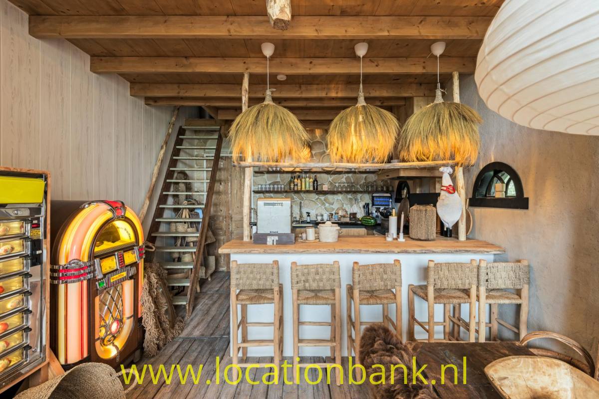 Holztheke mit hohen Hockern, drei geflochtenen Hängeleuchten und einer bunten Jukebox unter Holzbalken.