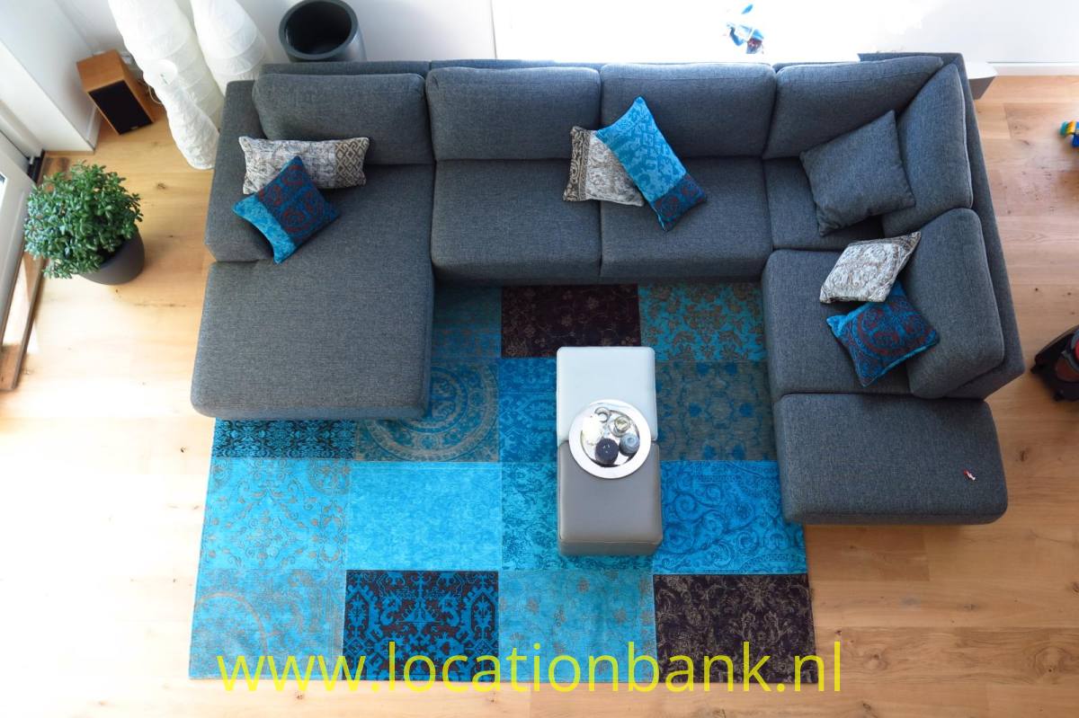 Ein graues Ecksofa steht um einen blauen Patchworkteppich mit einem hellen Couchtisch.