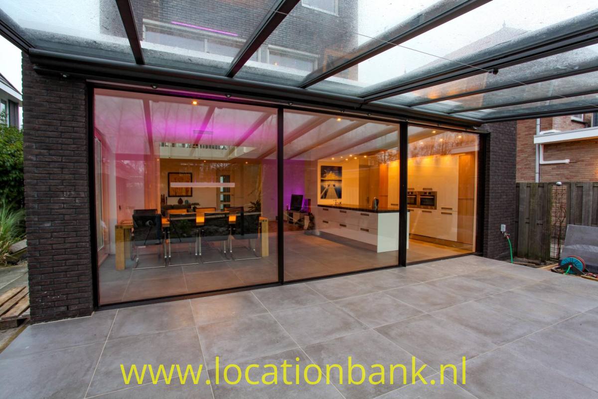 Verglaste Erweiterung mit transparentem Dach, sichtbarer Küche und Essecke, angrenzend an eine gepflasterte Terrasse.