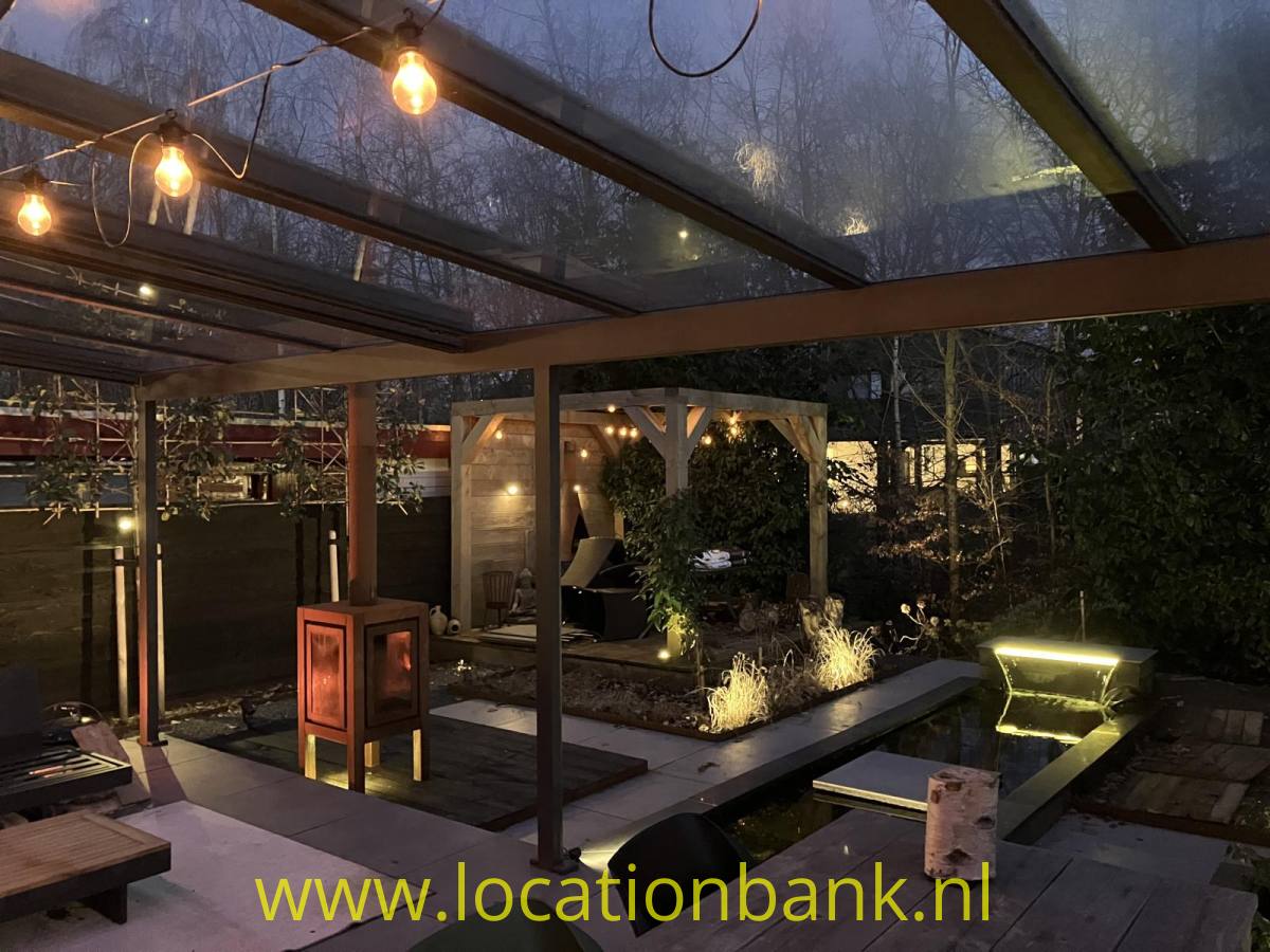 Überdachte Terrasse mit Lichterketten, einem Teich und beleuchteten Pflanzen in einem Garten.