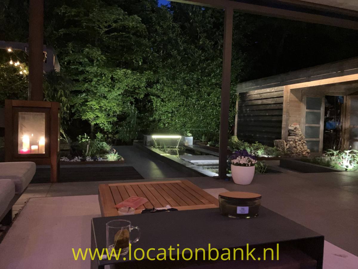 Überdachte Terrasse mit einem niedrigen Tisch, Gläsern und einem beleuchteten Garten mit einem langgestreckten Teich.