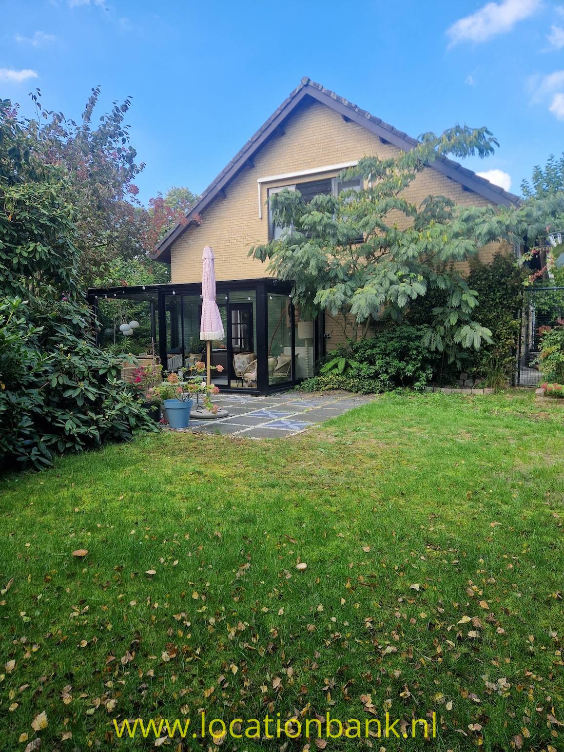 Haus mit steilem Satteldach und verglastem Anbau, umgeben von einem grünen Garten.