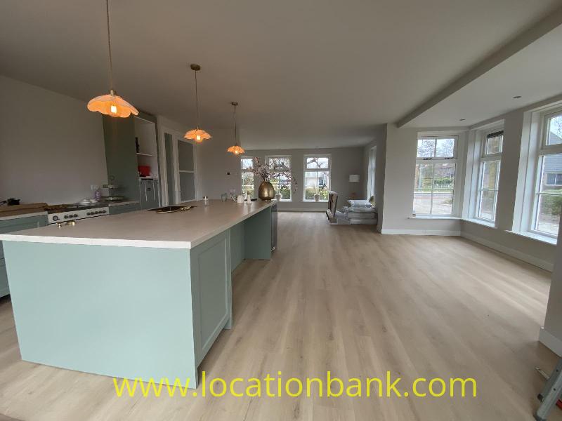 Open-plan living space with a kitchen island, pendant lights, wooden flooring and large windows.