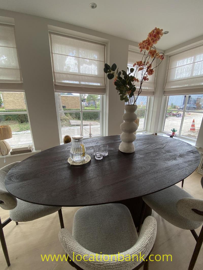 Oval dark dining table with a vase of branches, glasses and chairs in front of tall windows.