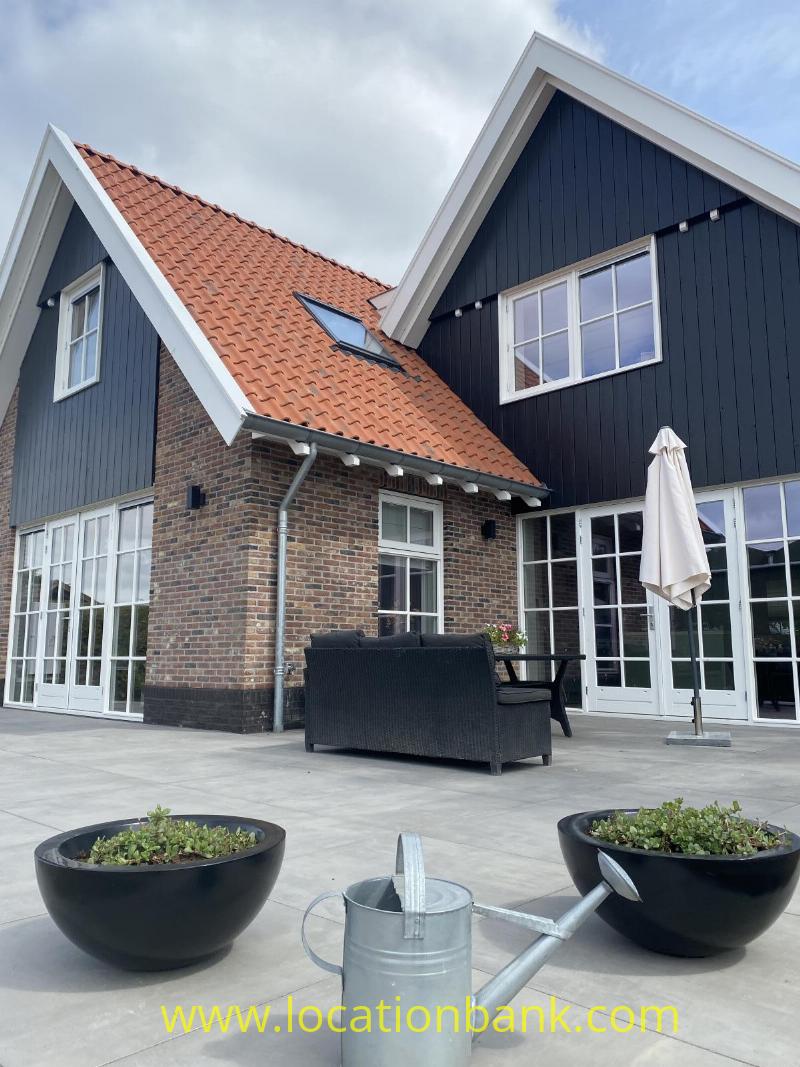 Dark façades with white frames, red roof tiles, a terrace and planters with a watering can in the foreground.