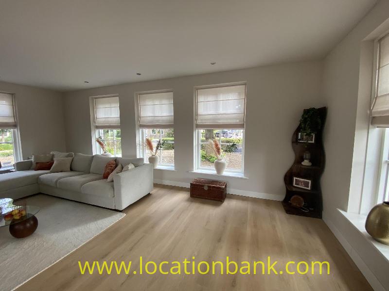 Living room with a light-coloured corner sofa, several tall windows and a dark open shelving unit.