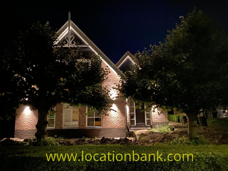 Illuminated brick house with gables, surrounded by trees and a low hedge beneath a dark night sky.
