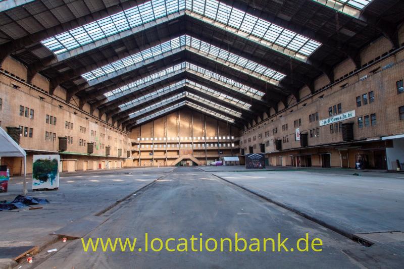 Große leere Halle mit gemauerten Seitenwänden, breiten Dachfenstern und einem Betonboden.