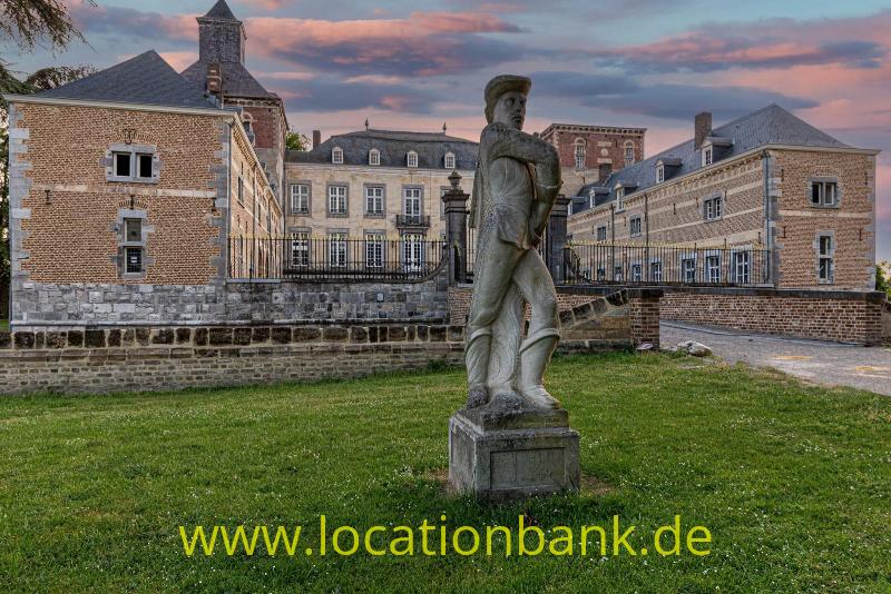 Ein steinernes Standbild steht auf einer Rasenfläche, hervor einem Schloss und einem Turm im Hintergrund.