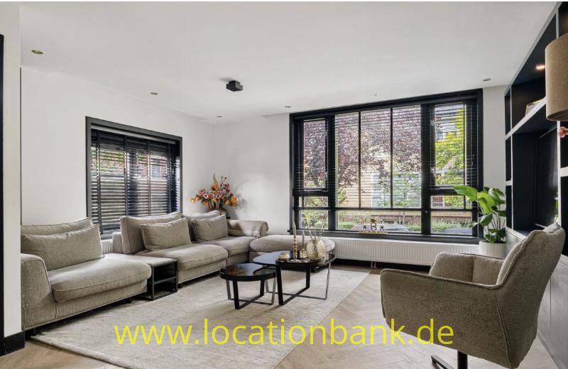 Helles Wohnzimmer mit beigefarbenen Sofas, einem Sessel, Couchtischen und großen Fenstern mit schwarzen Jalousien.