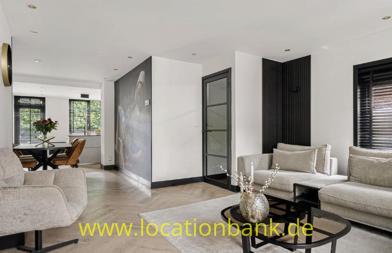 Modernes Wohnzimmer mit beigefarbenen Sofas, rundem Couchtisch, Holzboden und großen Fenstern mit Jalousien.