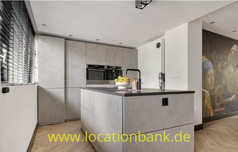 Graue Küche mit zentraler Kücheninsel, Einbaugeräten, einer Obstschale und einem großen Gemälde an der rechten Wand.