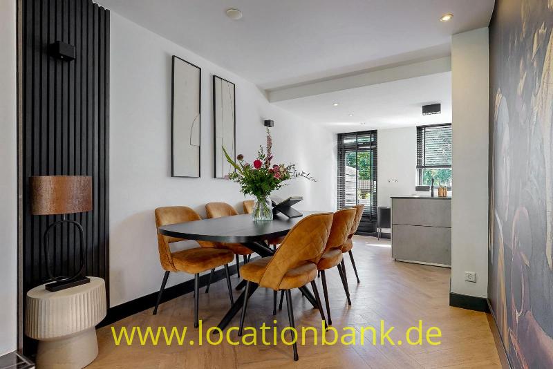 Ein schwarzer Esstisch mit einem Blumenarrangement und ockerfarbenen Stühlen steht vor einer schwarzen Lamellenwand.