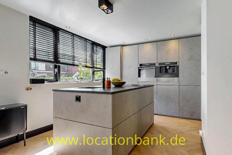 Küche mit einer zentralen Koch Insel, hohen Schränken, eingebauten Backöfen und horizontalen Jalousien vor den Fenstern.