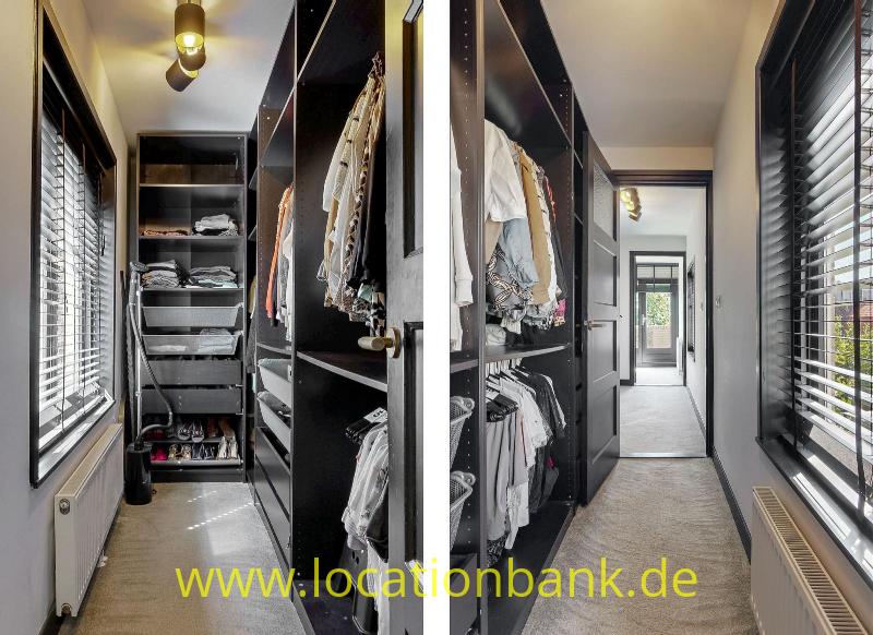 Ein Ankleidezimmer mit offenen schwarzen Schränken voller Kleidung, Fenstern mit Jalousien und einem langen Durchgang.