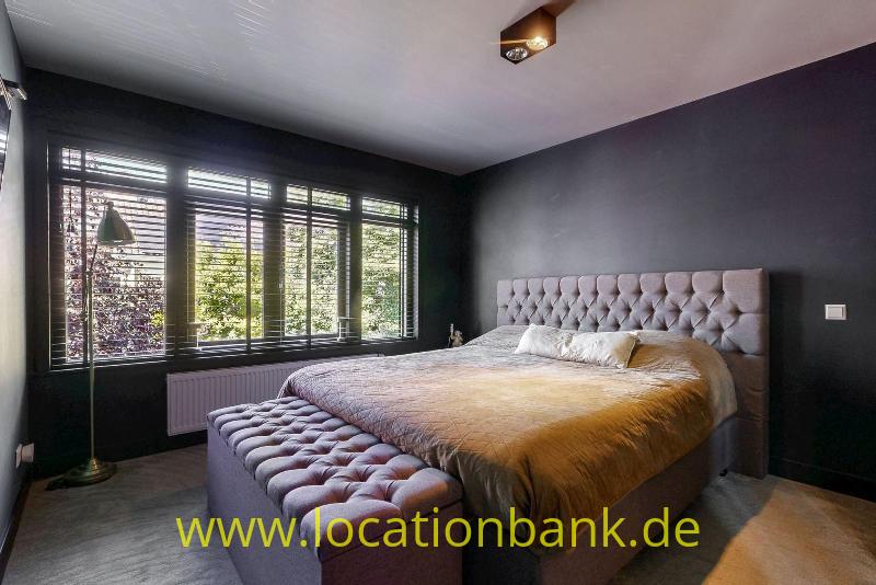 Schlafzimmer mit einem großen Bett, einem kapitonierten Kopfteil, dunklen Wänden und breiten Fenstern mit Jalousien.