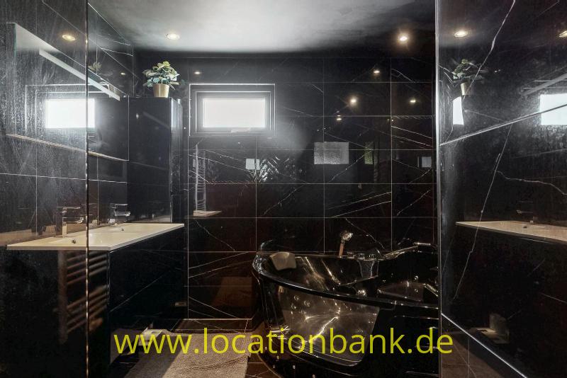 Eine freistehende schwarze Badewanne steht in einem Badezimmer mit schwarzen Fliesen, Spiegeln, einem Waschbecken und einem kleinen Fenster.