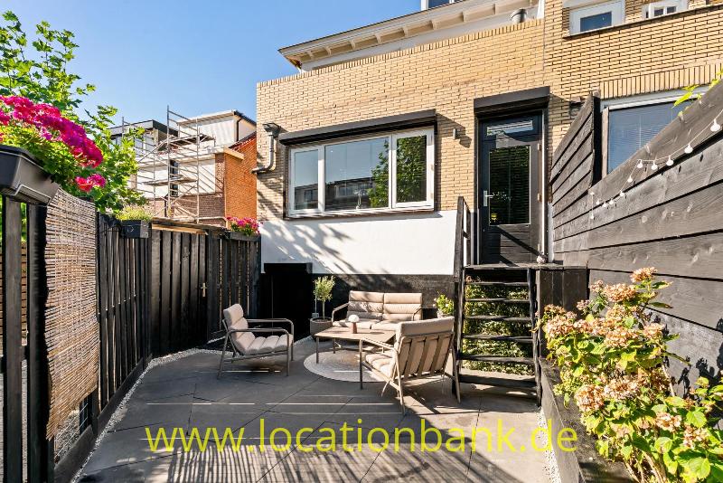 Gepflasterte Terrasse mit Sitzbereich, blühenden Pflanzen, einer Backsteinfassade und Stufen.