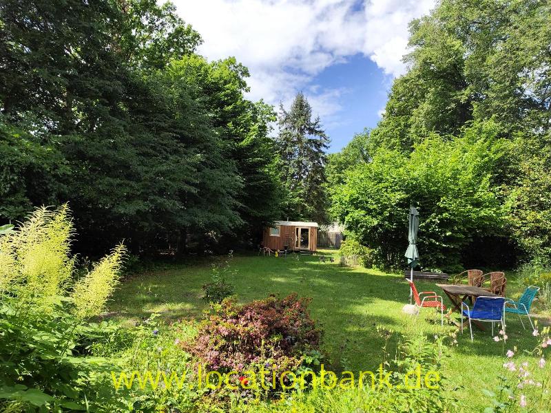 Ein grüner Garten mit hohen Bäumen, blühenden Pflanzen, einem hölzernen Gartenhaus und einer Sitzecke mit Sonnenschirm.