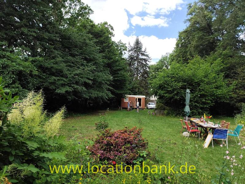 Grüner Garten mit einer Rasenfläche, Blumenbeeten, Gartenmöbeln und eines kleines Holzhauschen zwischen den Bäumen.