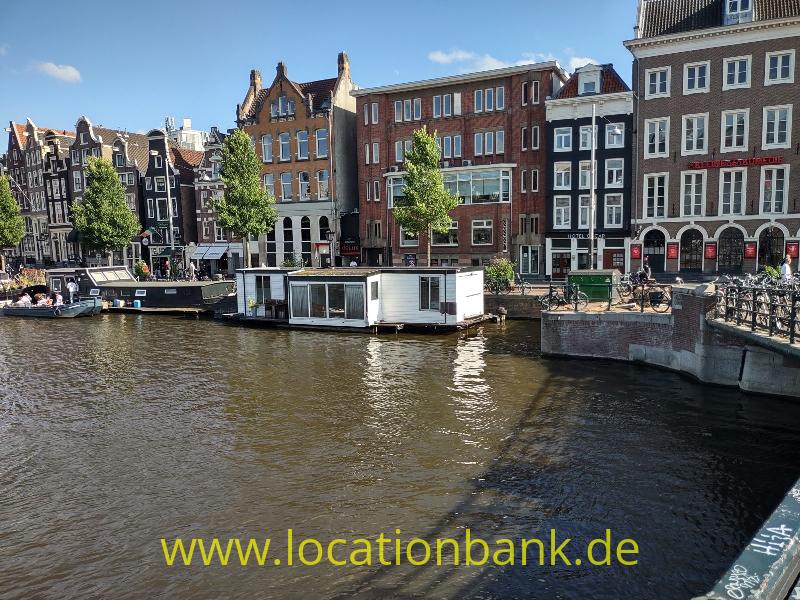 Ein Hausboot liegt in einem Kanal, dahinter stehen schmale hohe amsterdam Häuser und eine steinerne Kaimauer.