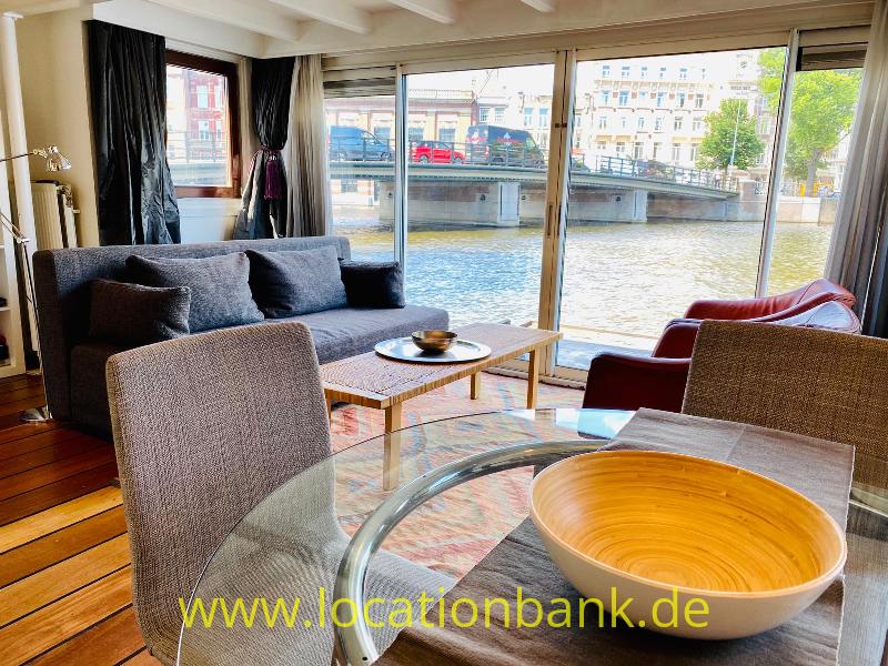 Wohnzimmer mit grauem Sofa und großen Fenstern mit Blick auf einen Amsterdam Kanal, eine Brücke und gegenüberliegende Grachten Häuser.
