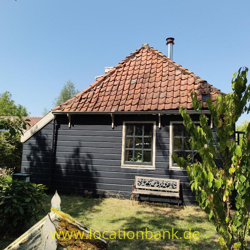 Schwarzes Holzhaus mit rotem Ziegeldach, zwei Fenstern und einer Bank im Garten.