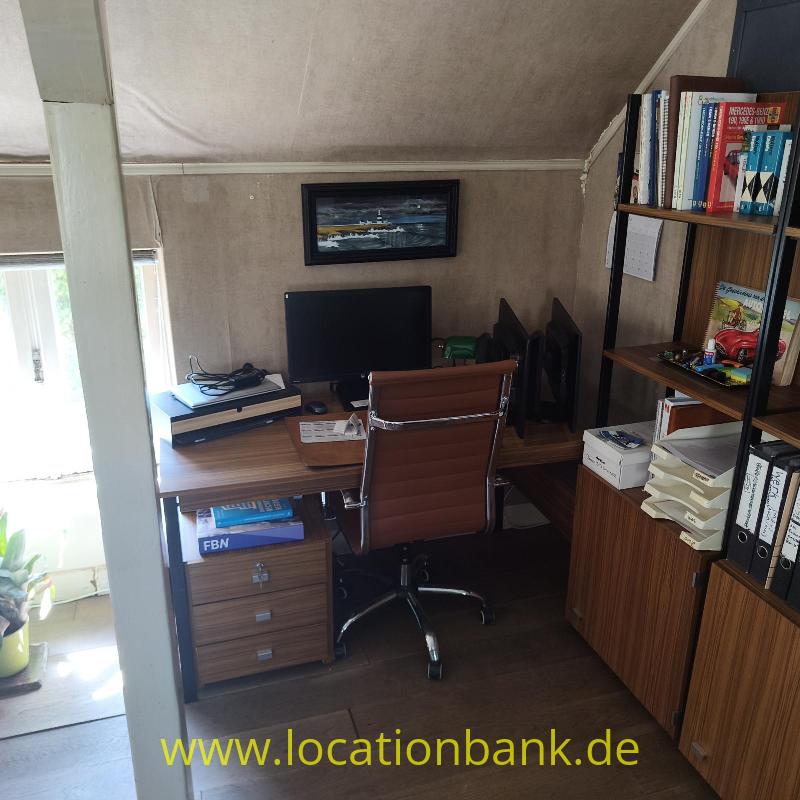 Ein Arbeitsplatz mit Schreibtisch, Computermonitor, Tastatur, Bürostuhl und einem mit Büchern gefüllten Regal.