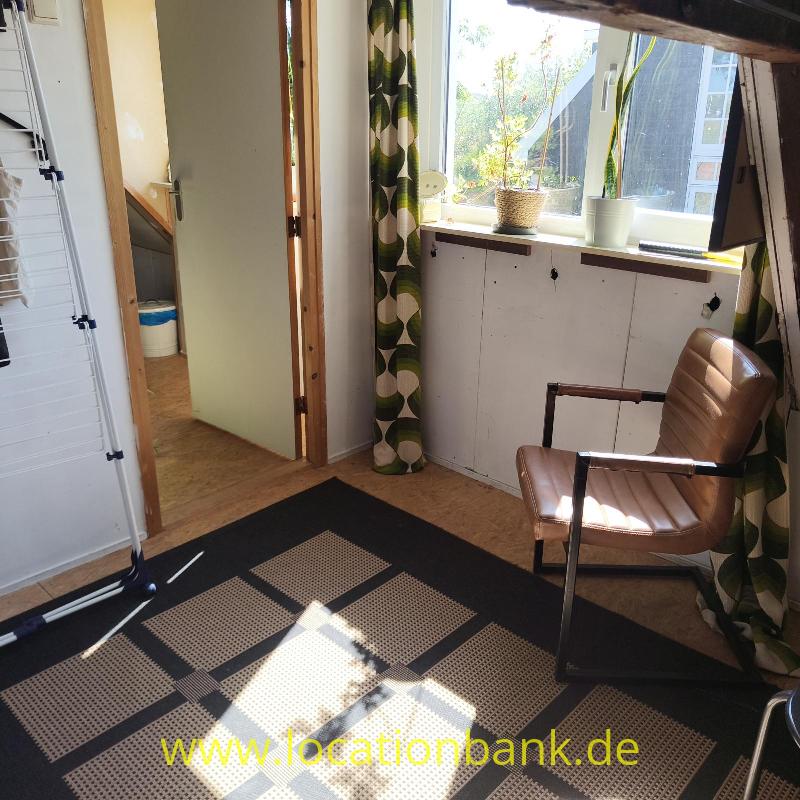 Brauner Sessel neben einem sonnigen Fenster mit Pflanzen, grünen Vorhängen und einem schwarzen Teppich mit geometrischem Muster.