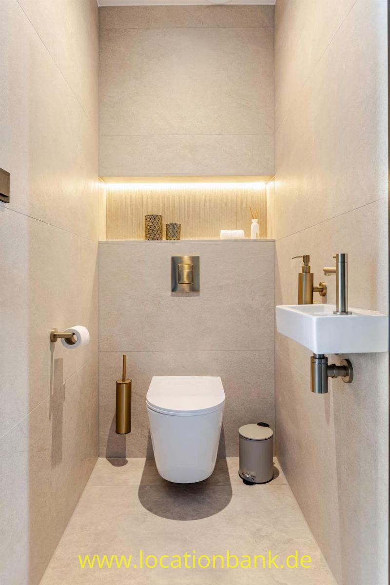 Beigefarbenes Badezimmer mit einer Toilette, einem Waschbecken, goldfarbenen Armaturen und einer Nische mit indirekter Beleuchtung.