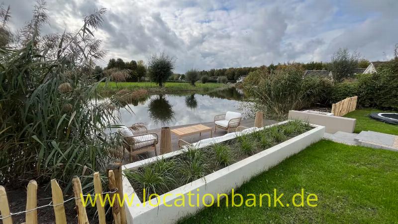 Rasen und erhöhte, weiß eingefasste Pflanzbeete an einem Teich sowie eine Holzterrasse mit Stühlen und hohem Schilf.