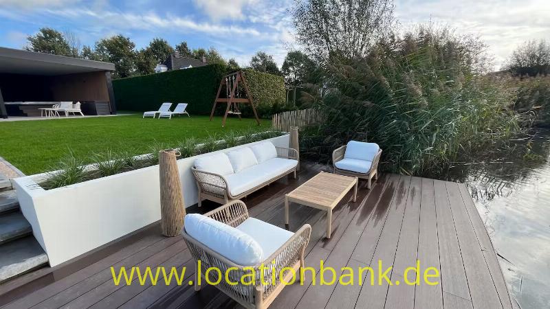Eine Holzterrasse mit einem geflochtenen Sofa, zwei Sesseln und einem niedrigen Tisch neben einem Teich.