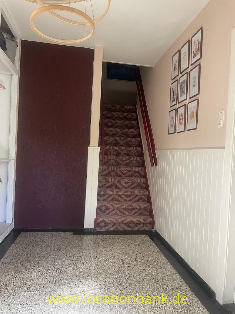 Eine Treppe mit gemustertem Läufer führt nach oben; an der rechten Wand hängen gerahmte Illustrationen.
