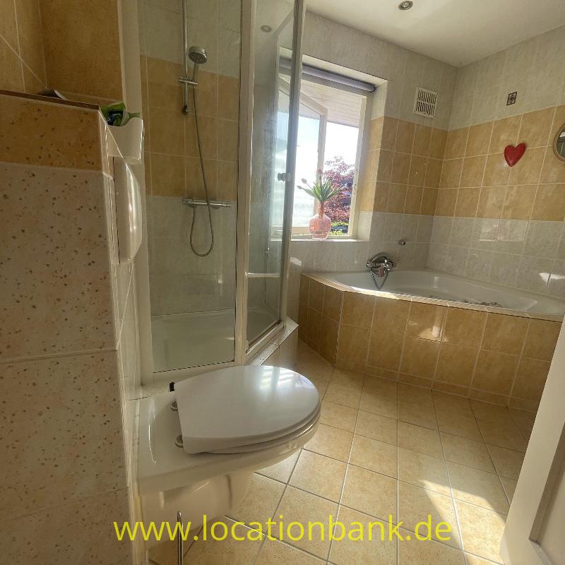 Gefliestes Badezimmer mit Duschkabine, Badewanne, Toilette und Fenster mit einer Pflanze.