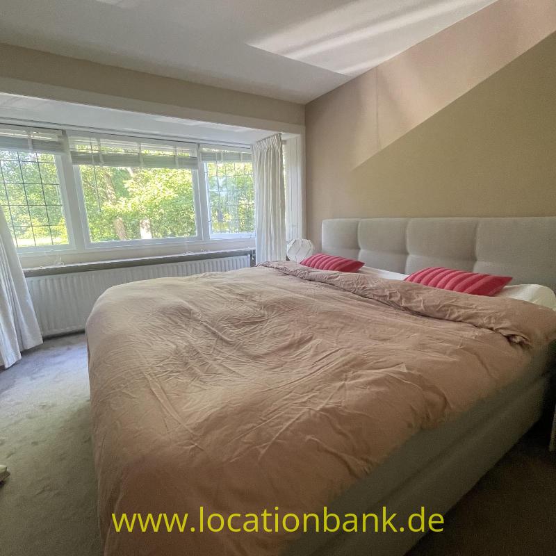 Ein großes Bett mit einer beigen Bettdecke, rosafarbenen Kissen und einem gepolsterten Kopfteil steht vor einem großen Fenster.