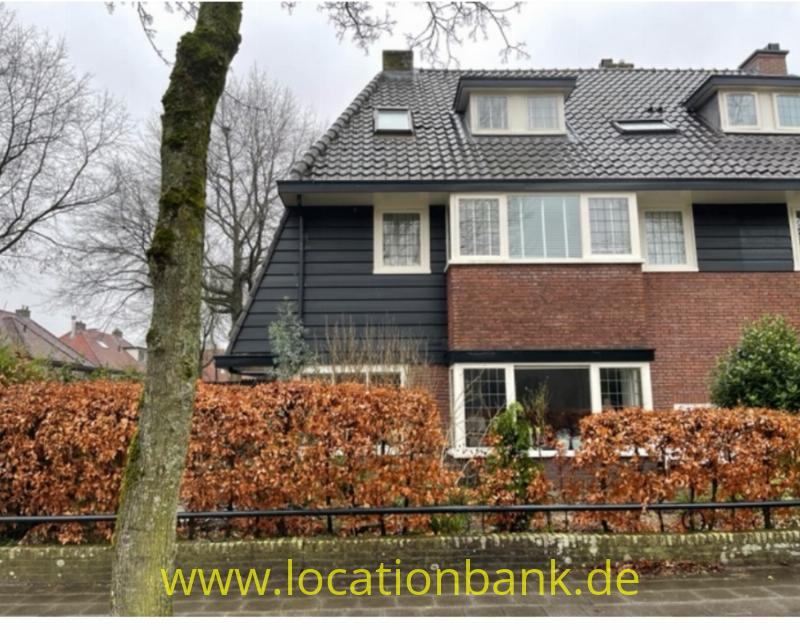 Haus mit dunkler Fassadenverkleidung, einer Backsteinfassade, Gauben, Dachfenstern, einer Hecke und einem Baum.