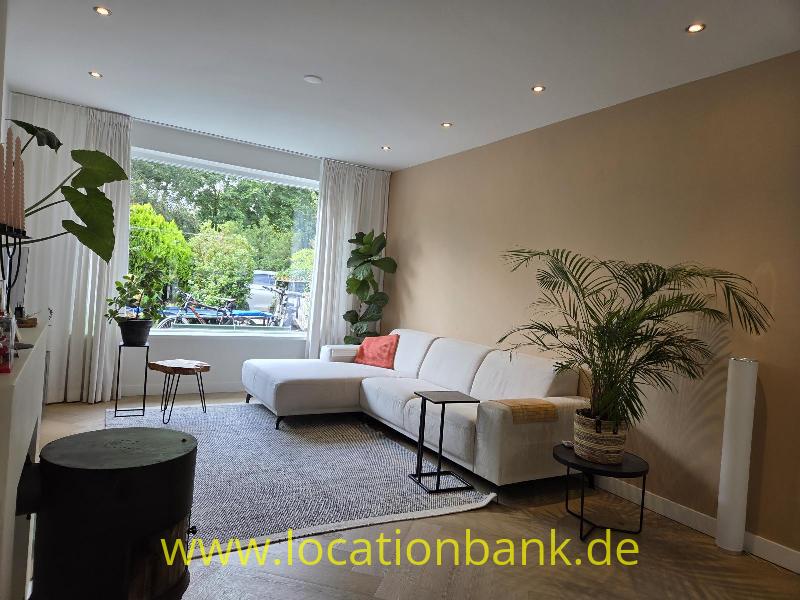 Wohnzimmer mit einem weißen Ecksofa, einem großen Fenster mit Vorhängen und grünen Pflanzen.