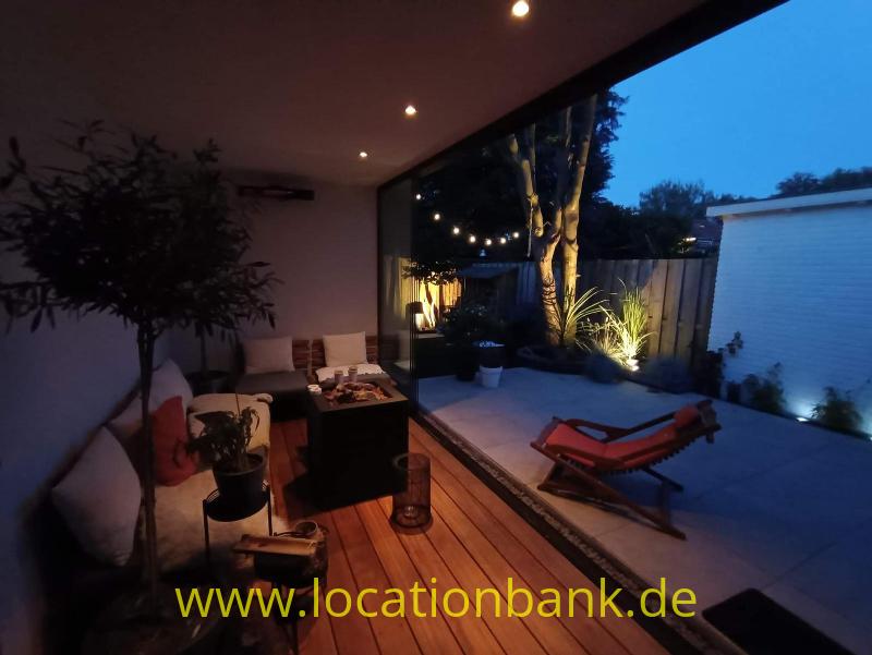 Überdachte Terrasse mit Sitzbereich, Holzdeck, Pflanzen und roten Liegestühlen neben einem gepflasterten Innenhof.