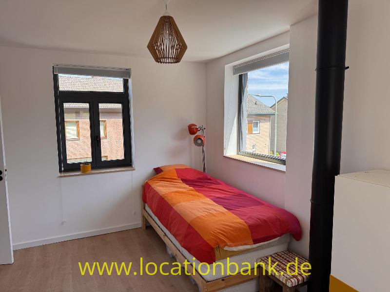 Ein Bett mit einer rot-orange-violett gestreiften Bettdecke steht zwischen zwei Fenstern und einem schwarzen Rohr.